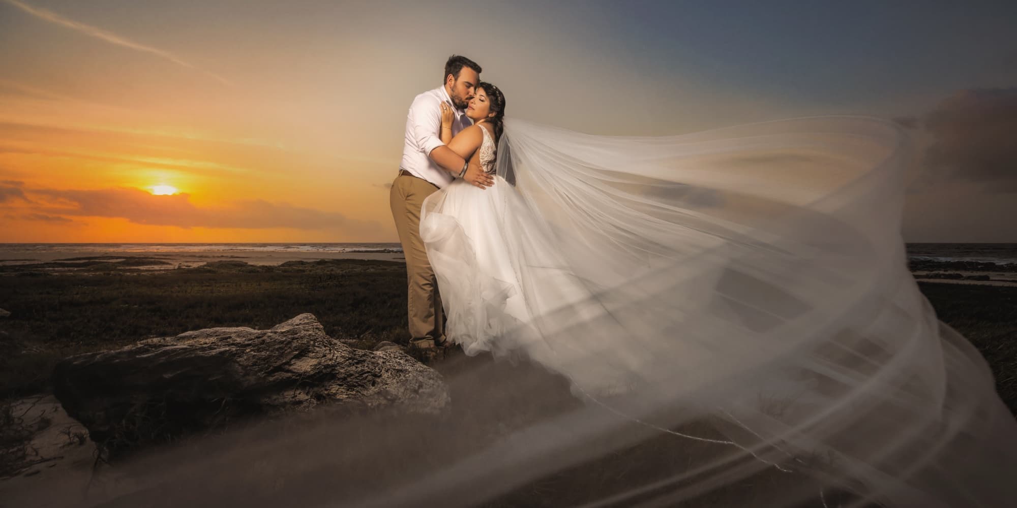 Nikole and Nick embracing at sunset on the Aruba coast with her cathedral veil streaming across the rocks and the sun dropping into the sea, photographed by Michael Anthony Photography