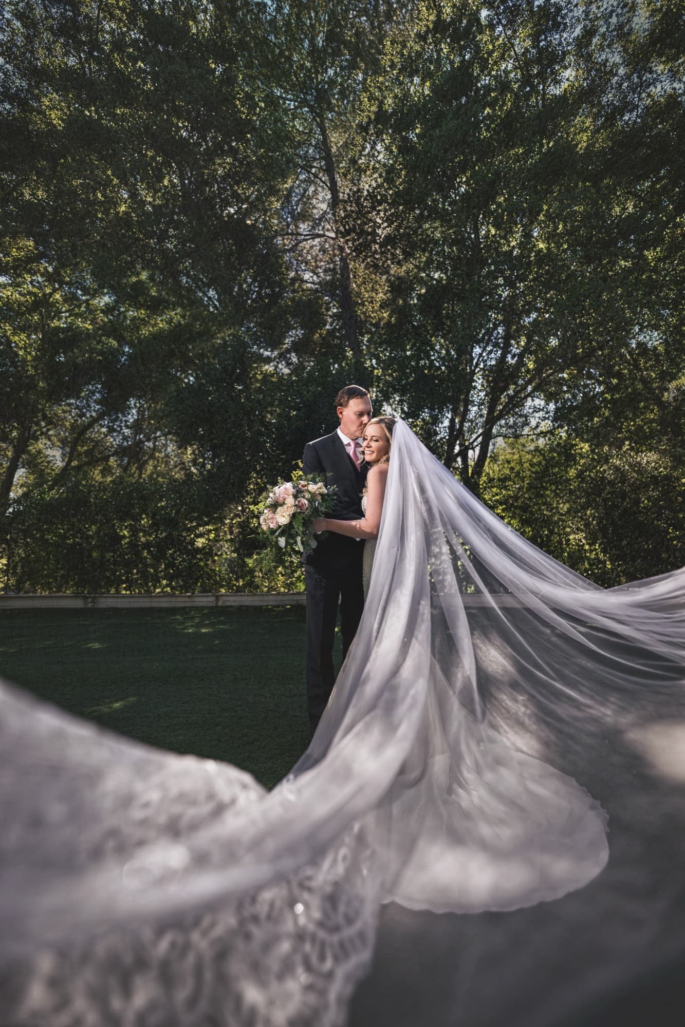 Monica and Colin beneath the oaks at Calamigos Ranch, her cathedral veil sweeping across the entire lawn in front of them, photographed by Michael Anthony Photography