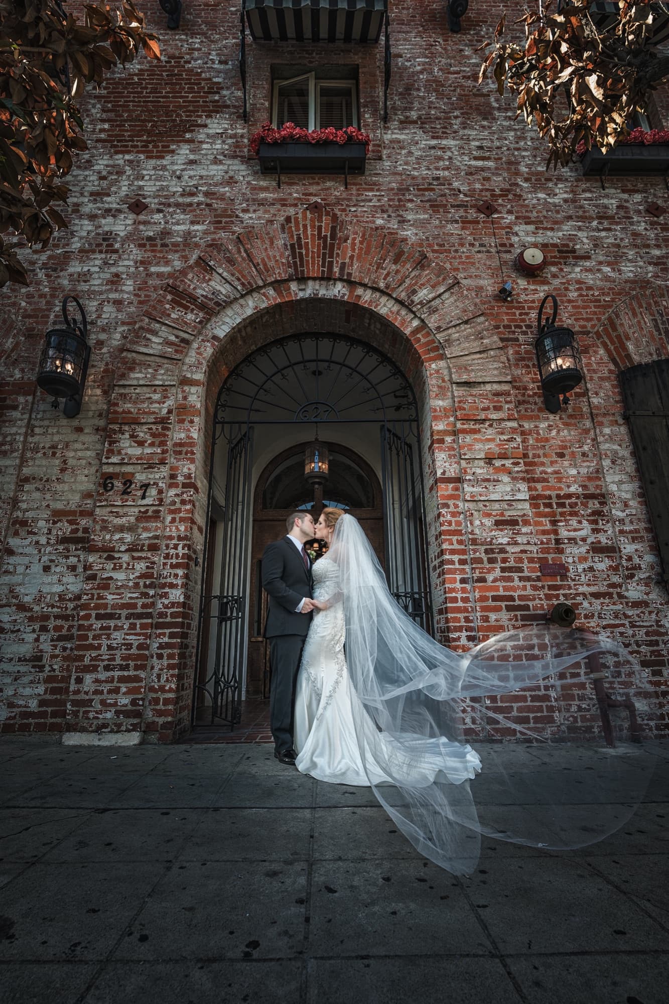 Briana and Peter kissing in the brick archway entrance of Carondelet House with her cathedral veil sweeping across the sidewalk, photographed by Michael Anthony Photography