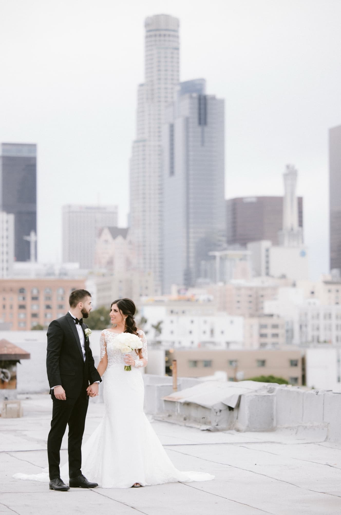 Hela and Yousef standing together on a downtown rooftop with the US Bank Tower and Los Angeles skyline rising behind them, photographed by Michael Anthony Photography