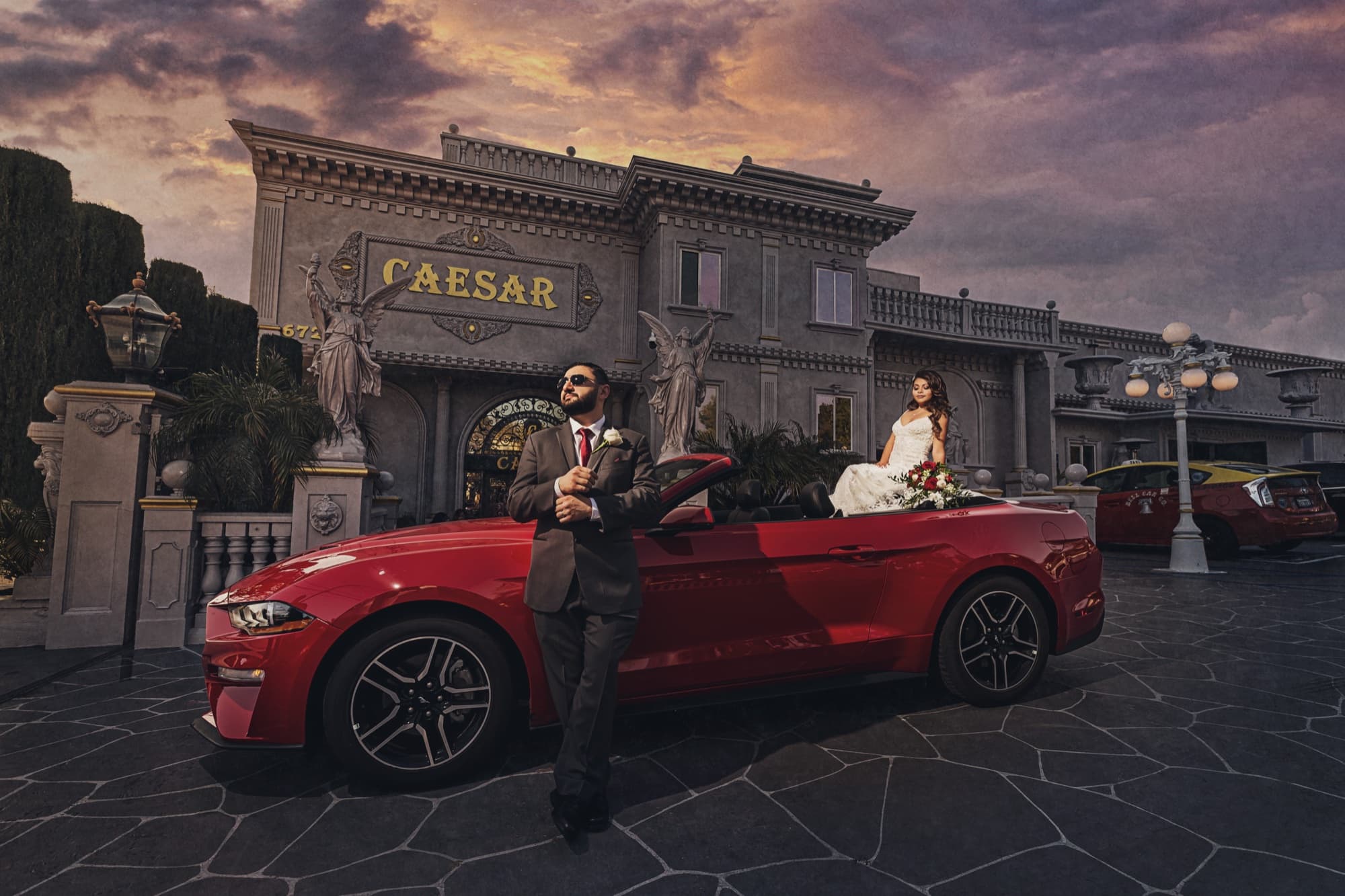 Angela seated in a red Mustang convertible and Arthur standing beside it in front of The Great Caesar's Banquet Hall under a dramatic evening sky, photographed by Michael Anthony Photography