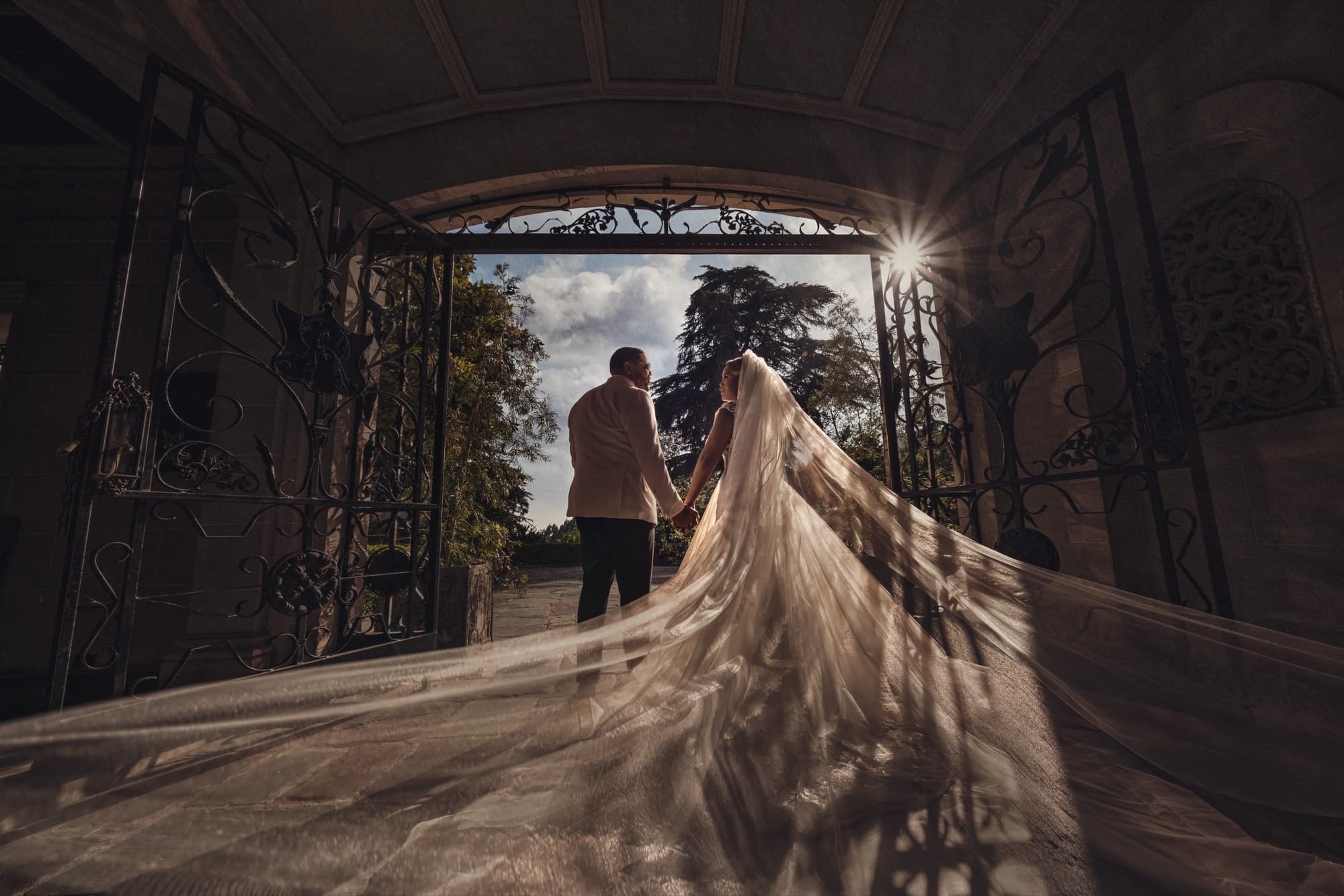 Shaniece's veil sweeping through Greystone Mansion's iron gate as she and Jarell walk into the sunlit colonnade, photographed by Michael Anthony Photography