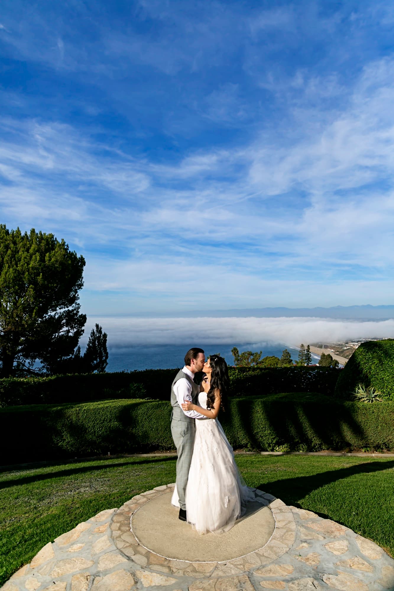 Stephanie and Josh kissing on the stone overlook at La Venta Inn with the marine layer rolling over the Palos Verdes coastline behind them, photographed by Michael Anthony Photography