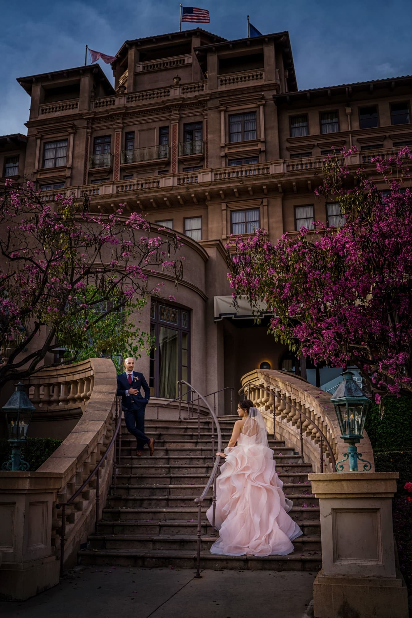 Ilene in a blush ruffled ballgown climbing the blossom-framed grand staircase of The Langham Huntington Pasadena at dusk, photographed by Michael Anthony Photography