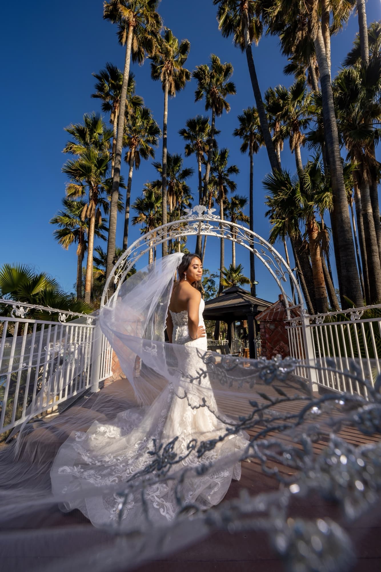 Karen beneath the white moon-gate arch at Rancho de las Palmas, her sparkling cathedral veil catching the flash against palms and a deep blue sky, photographed by Michael Anthony Photography