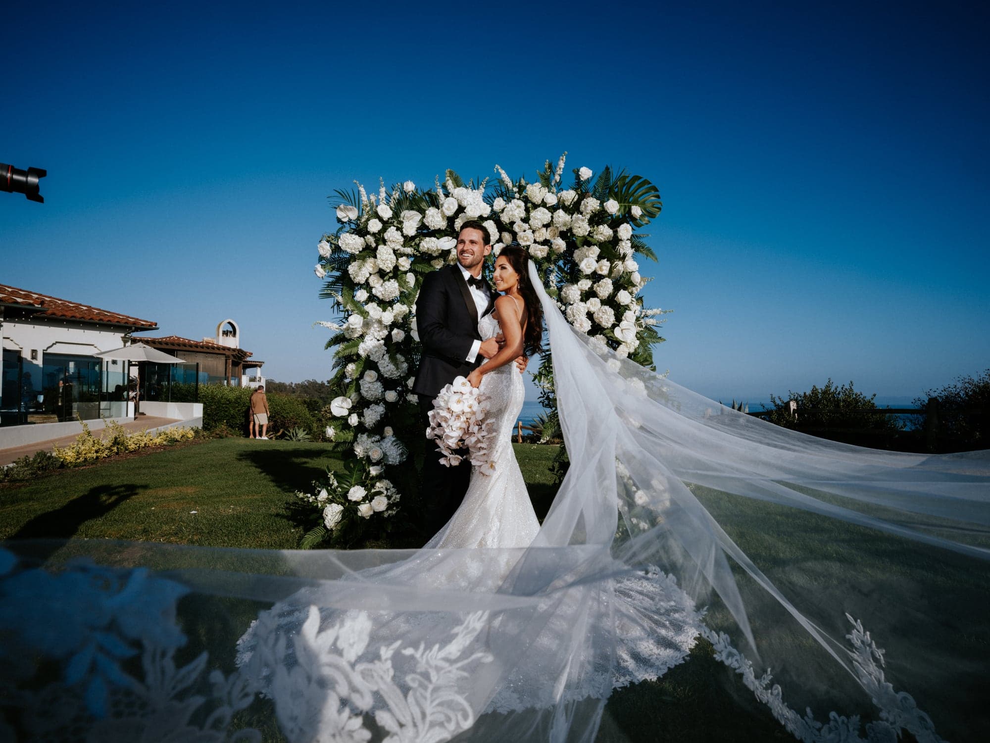 Tiana and Griffith beneath a white floral arch overlooking the Santa Barbara coast