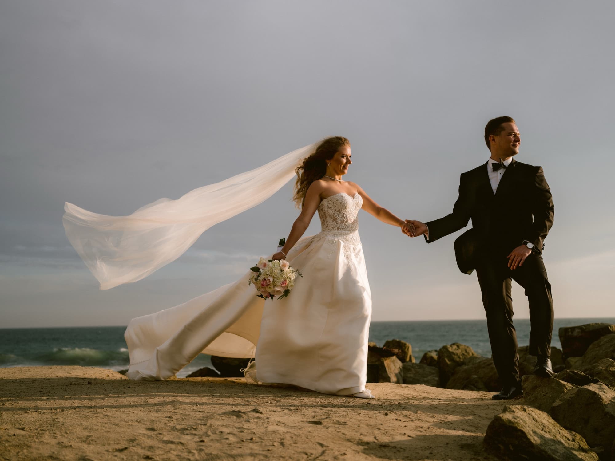 Anna and Alexey holding hands beside the Pacific after their Ritz-Carlton Laguna Niguel wedding, her veil and satin skirt lifted by the coastal wind, photographed by Michael Anthony Photography