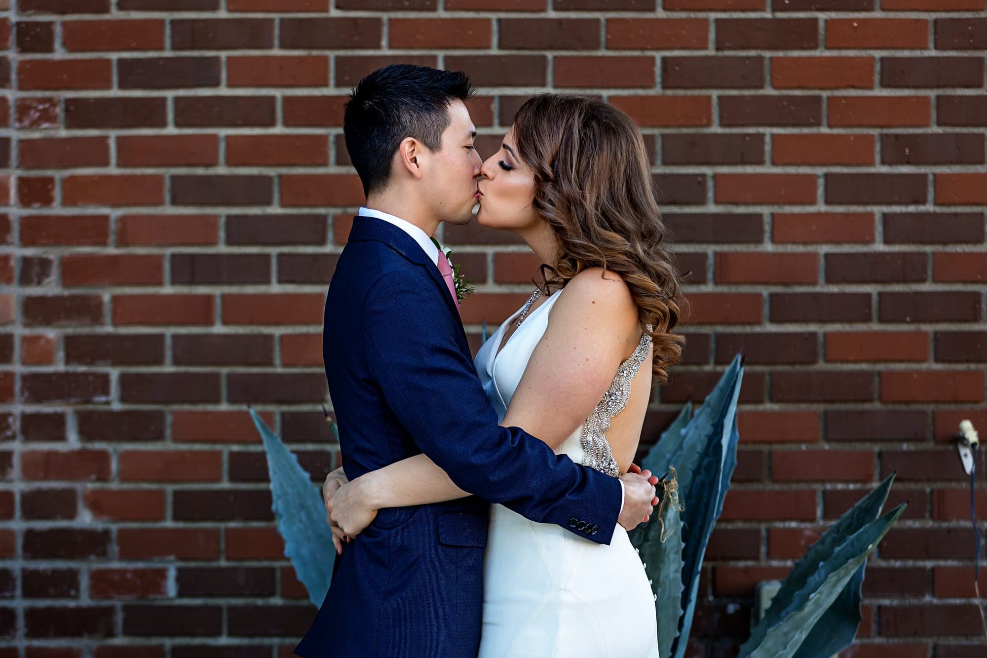 The Kaijoka newlyweds sharing a kiss against a brick wall at Vertigo Event Venue