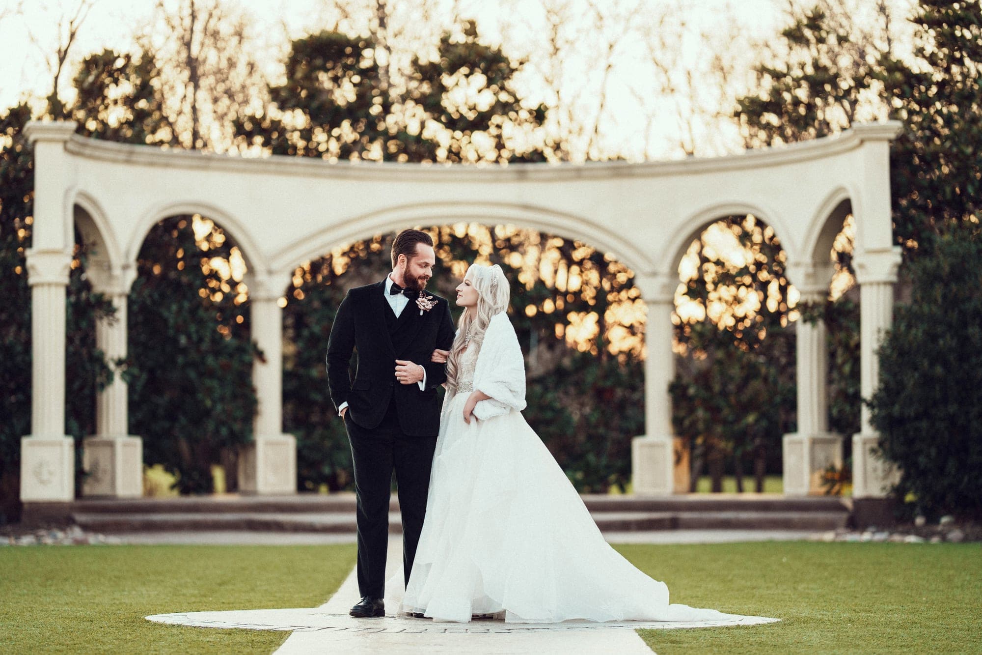 Carly and Christopher beneath the garden colonnade at Knotting Hill Place at sunset, her white fur wrap against his black velvet tuxedo