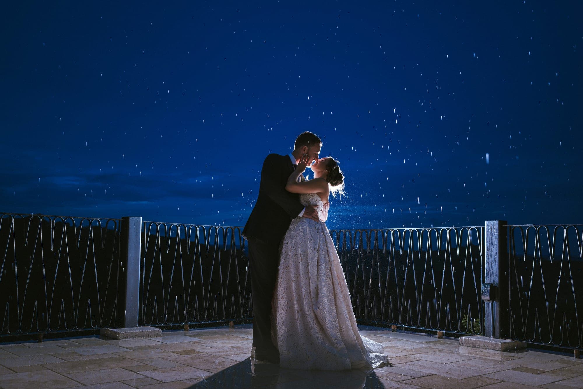 Kelsey and Eric kissing at night in backlit rain on the Villa Antonia terrace, the sky deep blue behind them