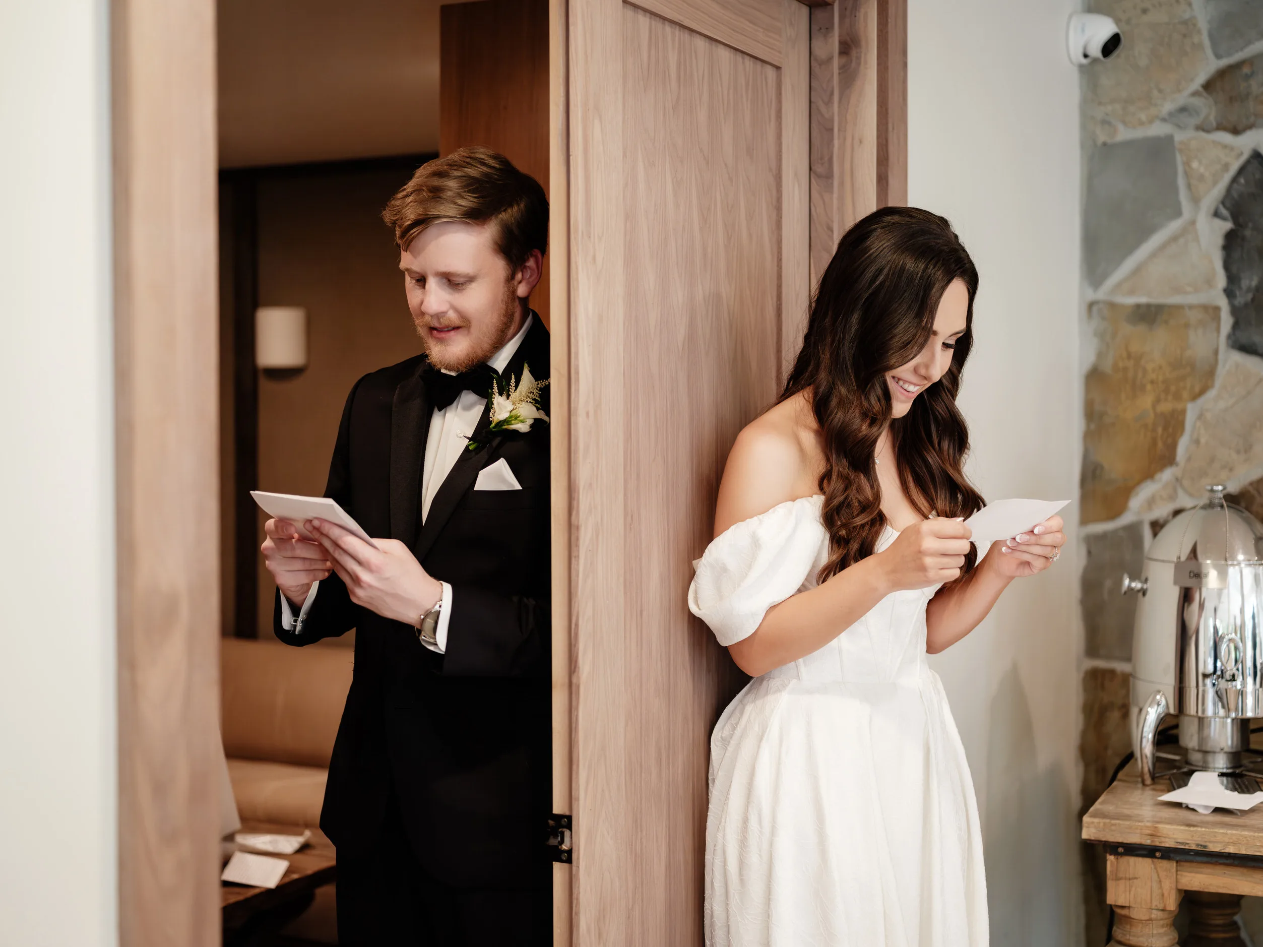 A groom in a black tuxedo and a bride in a white robe stand apart reading letters, smiling, in the Birchwood Room with stone and wood decor.