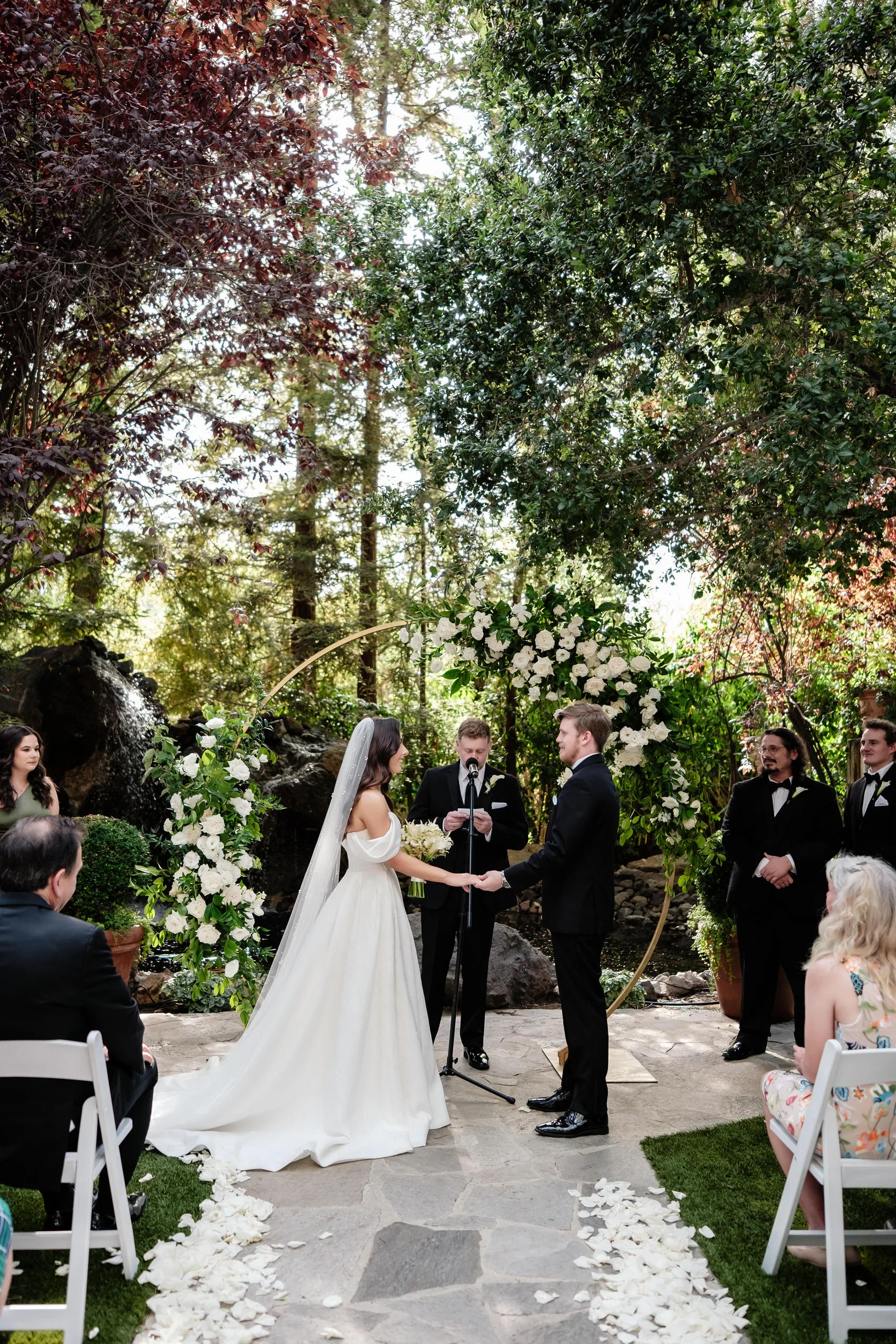 A bride and groom hold hands during their outdoor wedding ceremony at Calamigos Ranch, surrounded by lush greenery and white flowers, with a small audience seated on both sides.