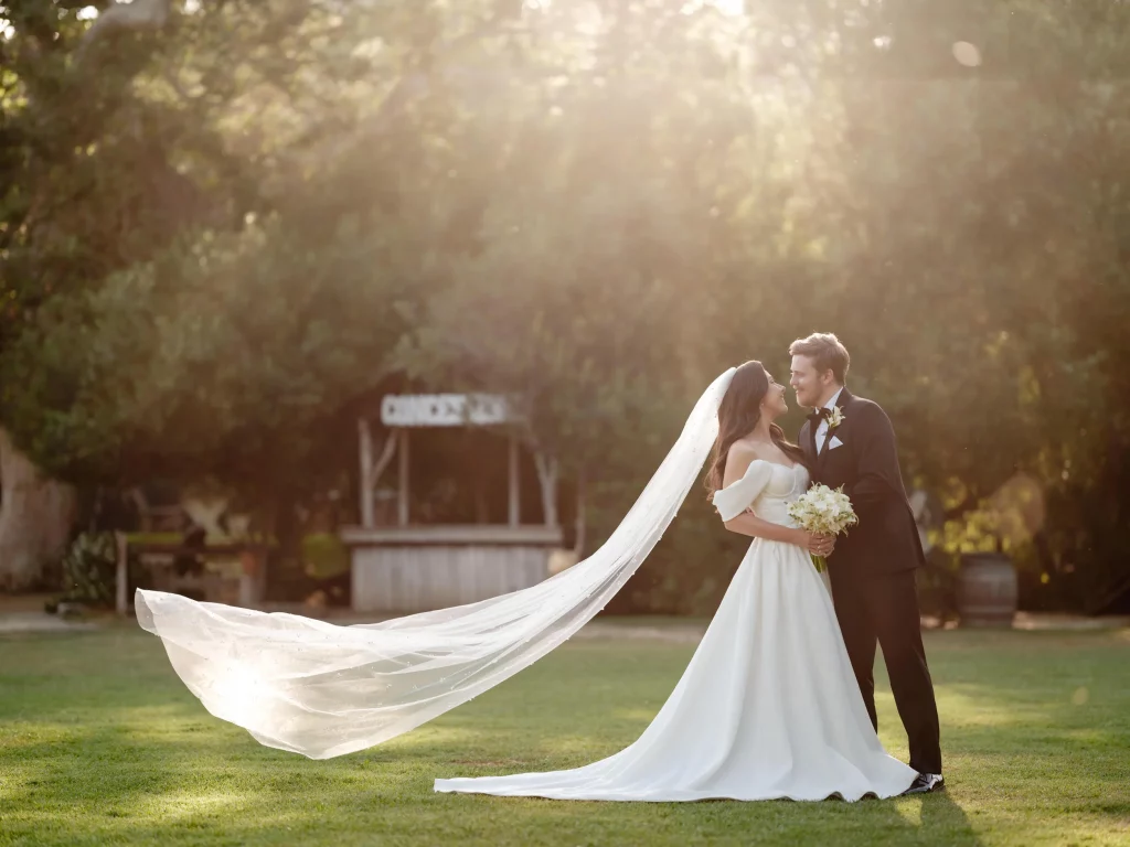 A newlywed couple sharing a tender moment outdoors at Calamigos Ranch. The bride, in a flowing white gown with a long veil, and the groom, in a black tuxedo, stand