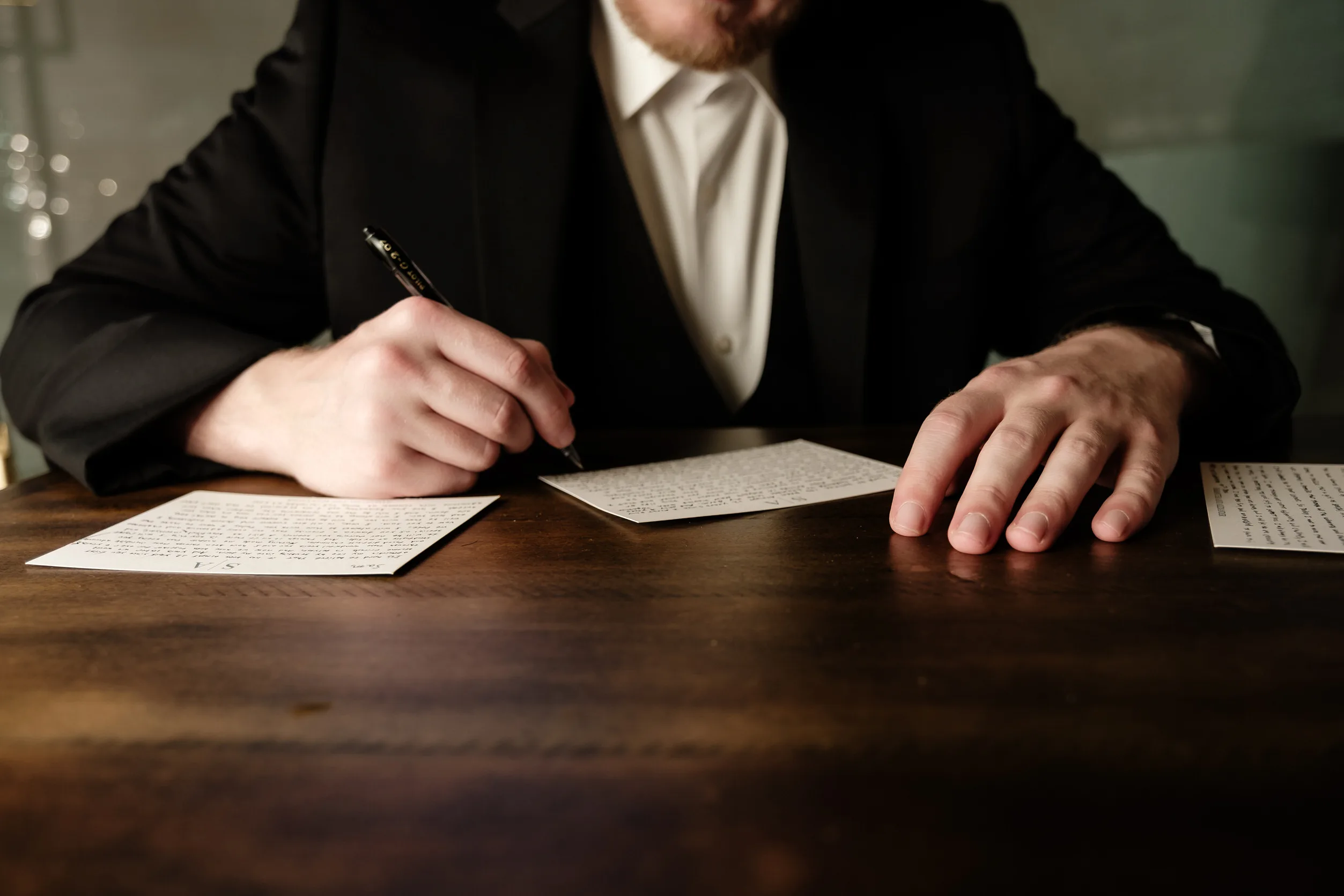 A man in a suit, visible from the chest down, sits at a wooden desk in the Birchwood Room writing on a paper with a pen, with another sheet of paper beside him.