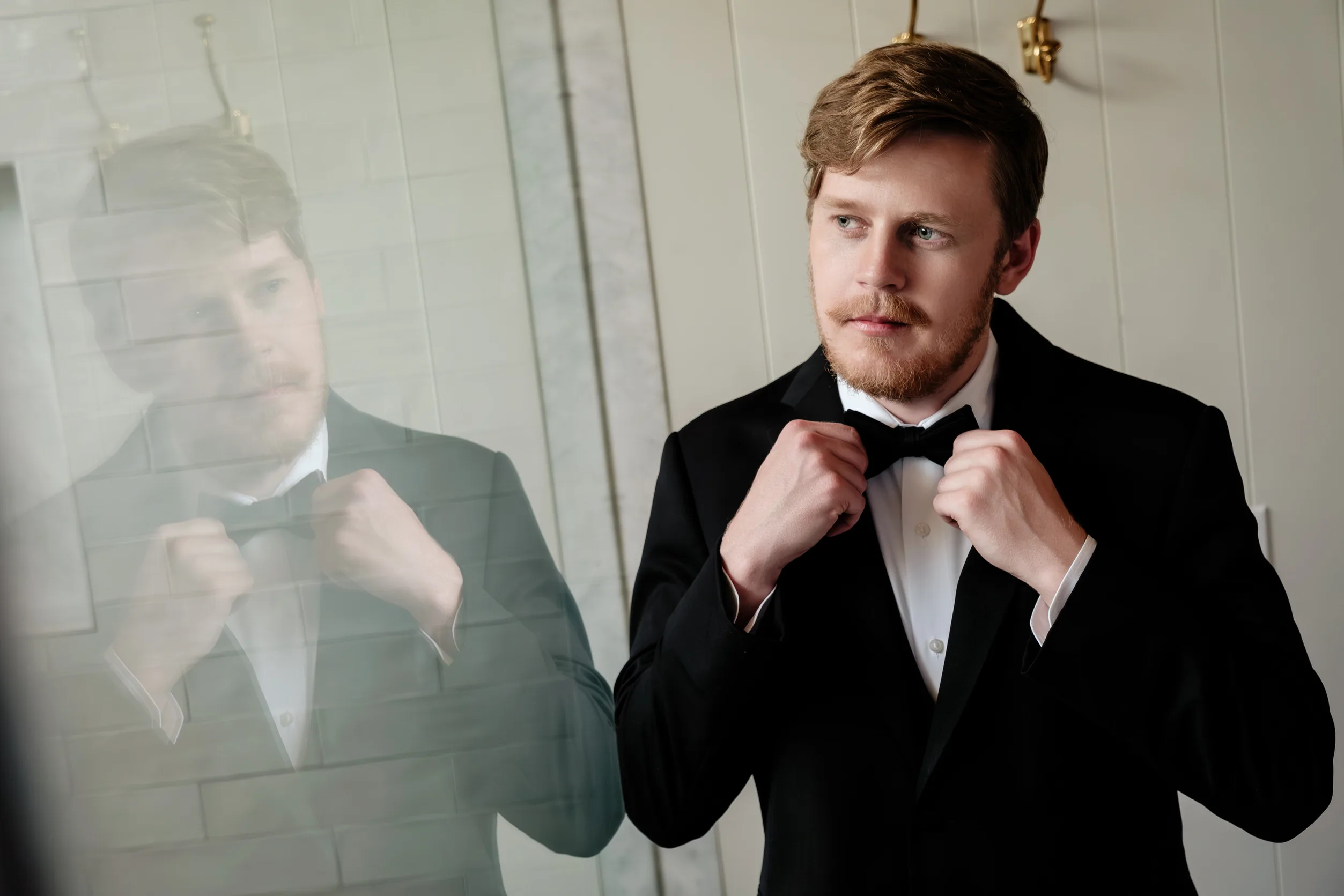 A man in a black suit and bow tie adjusts his tie while looking thoughtfully at his reflection in a mirror, in a room with elegant decor at a Calamigos Ranch Wedding.