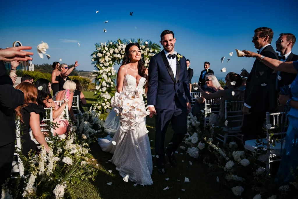Tiana & Griffith, a joyful bride and groom, walk hand-in-hand down the aisle as guests throw flower petals on a sunny day with a clear blue sky at their Ritz Carlton Santa Barbara Wedding