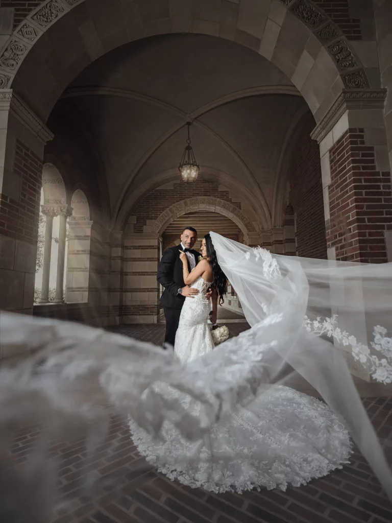 A newlywed couple embraces under an arched stone corridor, with the bride's long, lace-detailed veil flowing towards the camera. soft lighting enhances the romantic atmosphere.